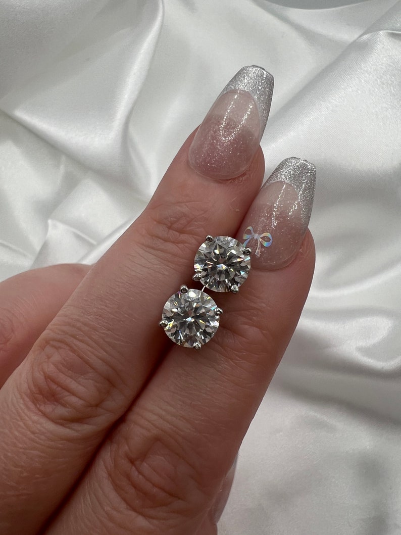 May include: A pair of round, brilliant-cut diamond stud earrings. The diamonds are set in a silver-colored metal and sparkle brightly. The earrings are displayed against a white, silky background.