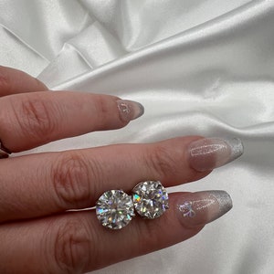May include: A pair of round, clear gemstone stud earrings. The gemstones are set in a silver or platinum setting. The earrings are displayed on a white satin background.
