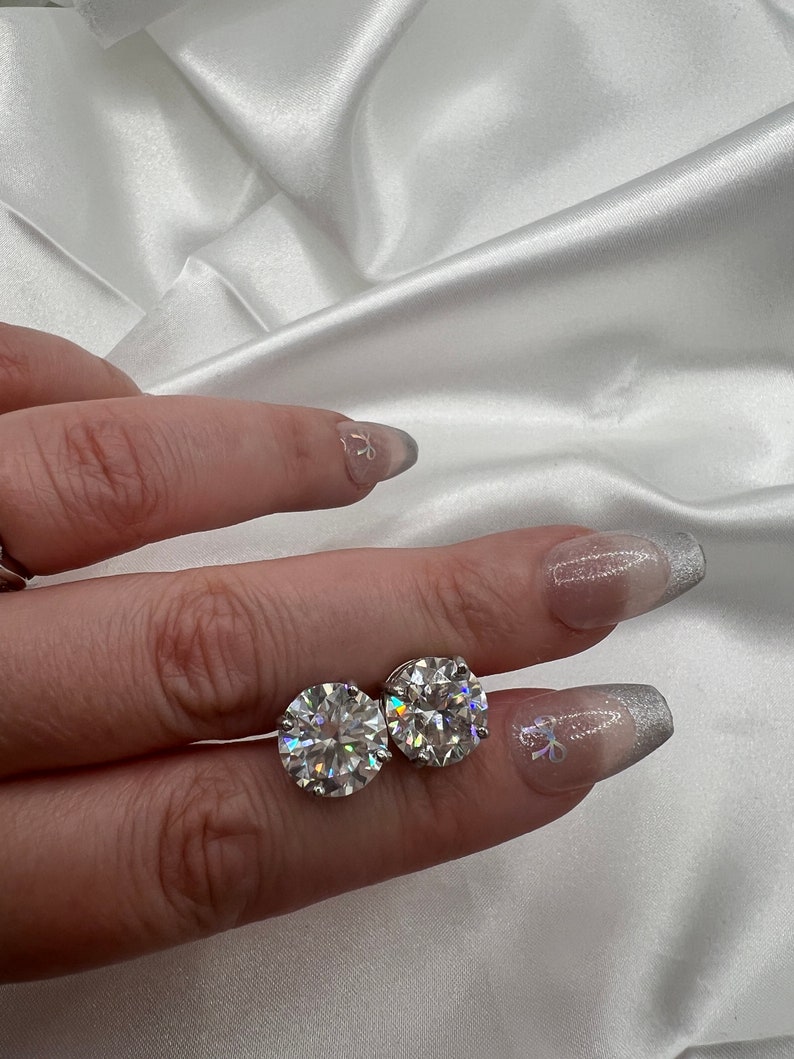 May include: A pair of round, clear gemstone stud earrings. The gemstones are set in a silver or platinum setting. The earrings are displayed on a white satin background.