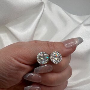 May include: A pair of sparkling, round-cut diamond stud earrings. The earrings are held against a backdrop of white satin fabric. The diamonds are clear and reflect light, creating a brilliant sparkle.