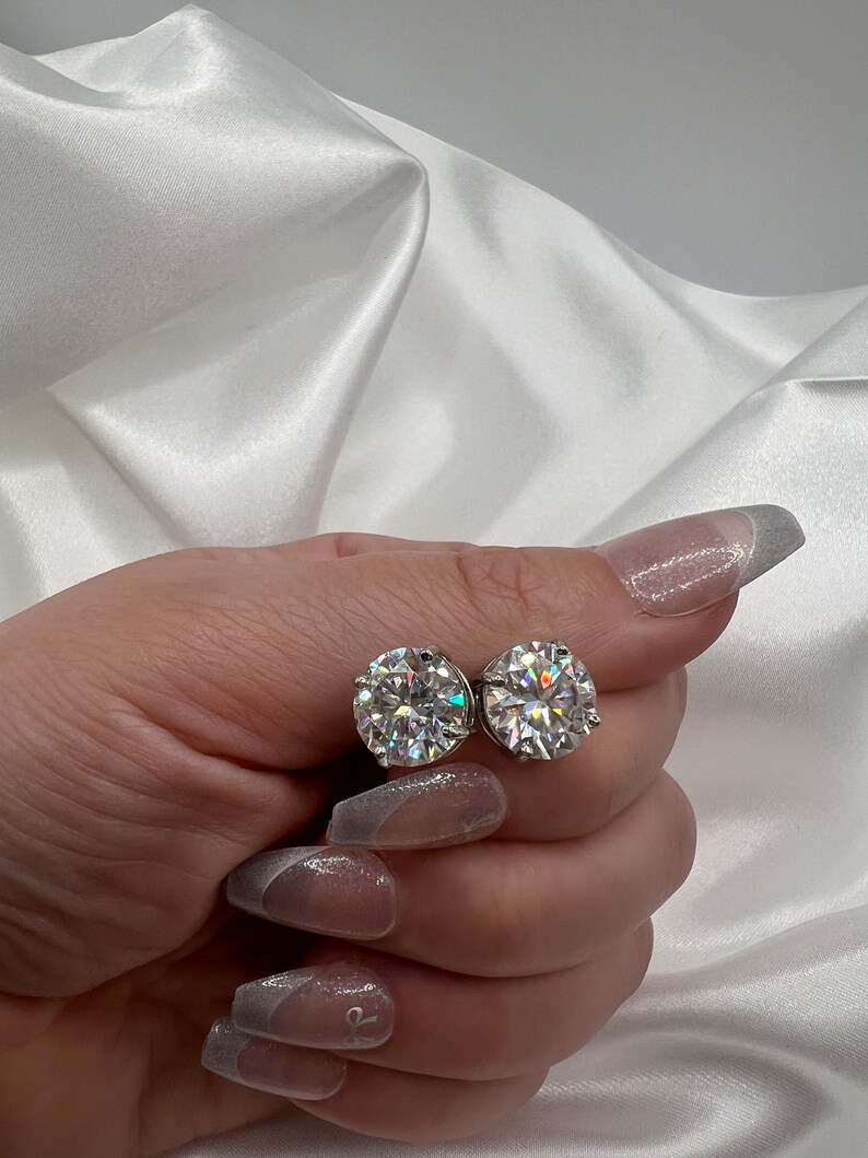 May include: A pair of sparkling, round-cut diamond stud earrings. The earrings are held against a backdrop of white satin fabric. The diamonds are clear and reflect light, creating a brilliant sparkle.