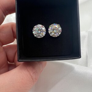 May include: Two round, sparkling diamond stud earrings displayed in a black jewelry box. The earrings are set in a silver or white metal and reflect light, creating a brilliant display. The box is held against a white satin background.