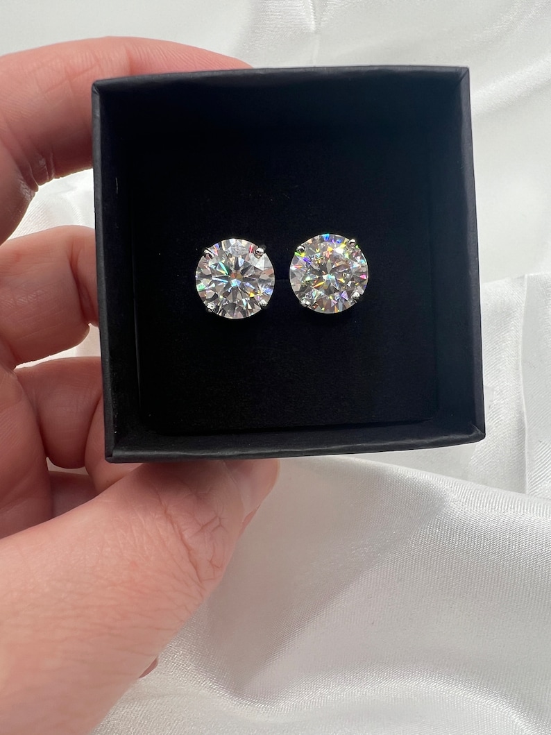May include: Two round, sparkling diamond stud earrings displayed in a black jewelry box. The earrings are set in a silver or white metal and reflect light, creating a brilliant display. The box is held against a white satin background.