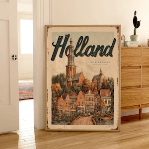 May include: A framed vintage-style poster with the word "Holland" in teal script. The artwork depicts a quaint European town with a church steeple, colorful buildings, and a bridge. The poster has a distressed, aged appearance.
