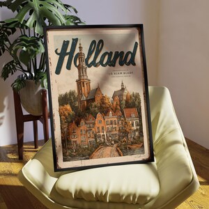 May include: A framed vintage-style poster with the word "Holland" in teal script. The artwork depicts a quaint European town with a church, bridge, and colorful buildings. The poster is in a black frame and rests on a cream-colored chair.