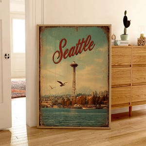May include: A framed vintage-style print featuring the Seattle skyline, including the Space Needle and waterfront. The word "Seattle" is written in red script across the top. The print has a distressed, aged appearance, with a blue and tan color palette.