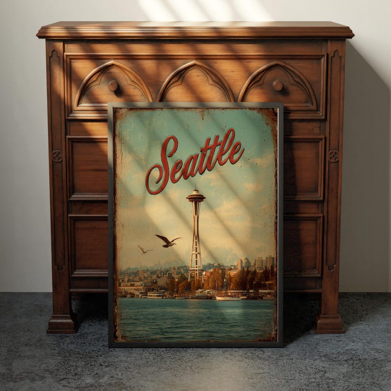 May include: A framed vintage-style Seattle travel poster featuring the Space Needle, birds, and waterfront. The poster has a distressed look with the word "Seattle" in red script. The poster is displayed in front of a wooden cabinet.