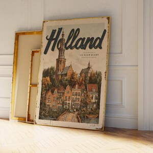 May include: A vintage-style poster with the word "Holland" in a dark blue script. The poster depicts a detailed illustration of a European town with a church, buildings, and a bridge. The poster is leaning against a white wall and a framed canvas.