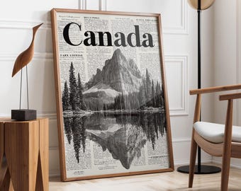 Puede incluir: Un grabado en blanco y negro enmarcado que representa un paisaje montañoso reflejado en el agua. La palabra "Canada" destaca en la parte superior. El grabado está en un marco de madera y se apoya en una pared blanca.