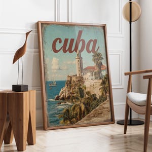 May include: A framed print featuring a vintage-style image of Cuba. The artwork depicts a coastal scene with a lighthouse, palm trees, and the word "cuba" in red script. The print is displayed in a room with wooden furniture.