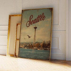 May include: A vintage-style framed print featuring the Seattle skyline, including the Space Needle and waterfront. The word "Seattle" is written in a cursive font. The print has a distressed, aged appearance, with a gold frame.