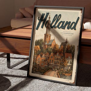 May include: Framed vintage-style poster with the word "Holland" in teal script. The artwork depicts a European town with a church, bridge, and colorful buildings. The poster is displayed on a wooden table.
