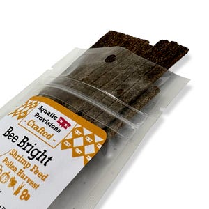 May include: A clear plastic package containing several dark brown, rectangular strips of Bee Bright shrimp feed and pollen harvest. The package has a white and orange label with the text "Aquatic Provisions - Crafted - Bee Bright".