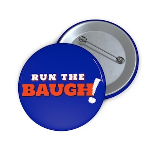 May include: A circular pin-back button with a royal blue background. The text RUN THE BAUGH! is printed in bold, orange, sans-serif capital letters across the center. The back of the button features a silver-toned metal pin mechanism.