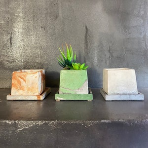 May include: Three square concrete planters in a row, each with a matching square base. The first planter is orange and white marbled, the second is green with a succulent, and the third is light gray. The planters are on a dark surface.