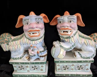 May include: A pair of colorful porcelain foo dogs, each with a playful expression and intricate details. The foo dogs are sitting on square bases with floral designs.