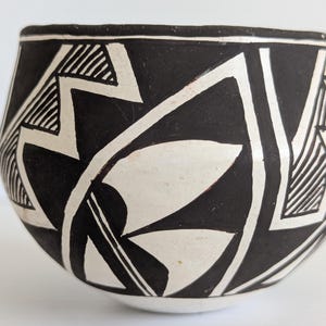 May include: A black and white ceramic bowl with a geometric design. The bowl features a rounded shape with a wide opening. The exterior is decorated with bold black and white patterns, including triangles, lines, and leaf-like shapes. The bowl is likely handmade.