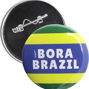 May include: A round button with a green, yellow, and blue striped design. The button has the text "Bora Brazil" in white letters.