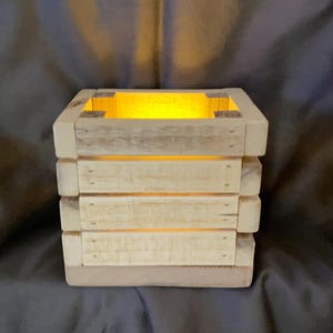 Puede incluir: Una caja de madera con un acabado de madera clara y un brillo cálido que emana del interior. La caja está hecha de varias piezas de madera apiladas, creando un estilo rústico y de granja.