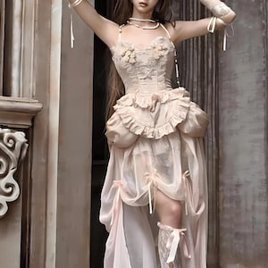 May include: A woman wearing a light pink, layered, ruffled dress with a lace bodice and a sheer overlay. The dress has a high neckline and a fitted bodice. The woman is standing in front of a stone wall.