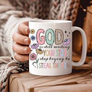 May include: White ceramic mug with a colorful floral design and the text "GOD is still writing YOUR STORY stop trying to STEAL THE PEN".