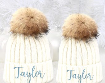 May include: Two white knit hats with faux fur pom poms. The hats have the name "Taylor" embroidered on the front in light blue thread.