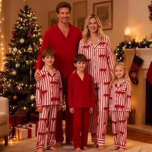 May include: A family of five wearing matching red and white striped pajamas with bow accents. The pajamas are paired with solid red sleepwear. A Christmas tree and fireplace are visible in the background, creating a festive holiday scene.