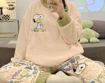 May include: A light pink Snoopy pajama set. The long-sleeved top features a Snoopy graphic and green trim on the collar and cuffs. The pajama pants have a white background with Snoopy graphics and various text designs. The pajamas are made of a soft, plush material.