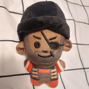 May include: A plush toy pirate with a black hat and eye patch. The toy has a brown face, ears, and arms, with an orange and brown outfit. The pirate has a stitched smile and is a soft, cuddly toy.