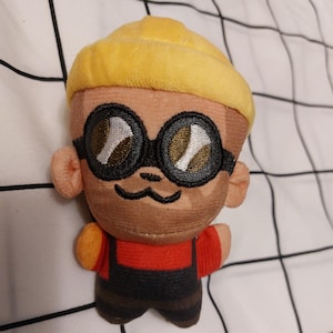 Op de afbeelding: Een pluche speelgoed van een personage met een gele muts, zwarte bril en een bruine snor. Het personage draagt een rood shirt en een donkerblauwe overall. Het speelgoed ligt op een witte stof met een zwart rasterpatroon.