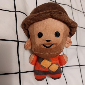 May include: A soft plush doll featuring a brown hat and beard, dressed in a red and brown patterned outfit. The doll has black eyes and a smiling face. The toy is made of soft fabric.