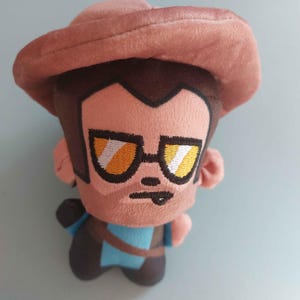 May include: A plush toy character wearing a wide-brimmed brown hat and yellow-tinted sunglasses. The figure has brown hair, a light skin tone, and a blue shirt with a brown strap across the chest. The facial features are embroidered with black thread.