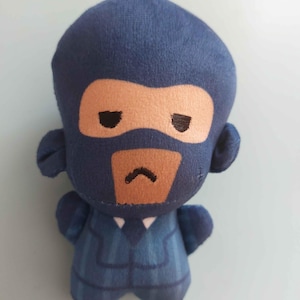 May include: A plush toy character wearing a dark blue pinstriped suit with a white shirt collar and a dark blue tie. The character has a dark blue balaclava mask covering the head, revealing a tan face area with black eyes and a small frown.