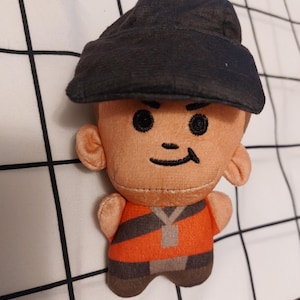 May include: A plush doll of a cartoon character wearing a black cap and an orange and grey striped shirt. The doll has a smiling face with black eyes and a black curved line for a mouth. The doll is made of soft fabric.