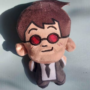 May include: A plush toy depicting a cartoon figure in a black suit, white shirt, and tie. The doll has brown hair, red-tinted glasses, and a neutral expression. The toy is made of soft, textured fabric.