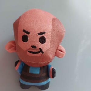 May include: A plush toy featuring a bald character with a peach-colored head, embroidered black eyes, eyebrows, and a mouth. The figure wears a blue and dark brown striped shirt with orange accents on the sleeves and dark brown pants.