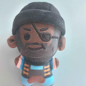May include: A small plush toy featuring a character with brown skin, a black beanie, and an eye patch over the left eye. The character has a dark beard, a blue and black outfit with orange accents, and pink stripes on the short sleeves.