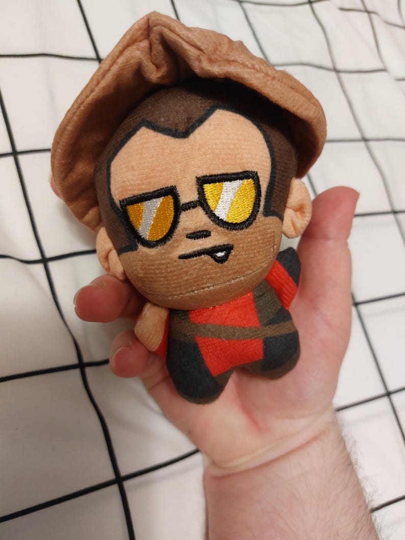 May include: A plush doll of a character wearing a brown hat and glasses with yellow lenses. The doll has a brown face, black eyebrows, and a small goatee. The doll is wearing a red and black outfit.