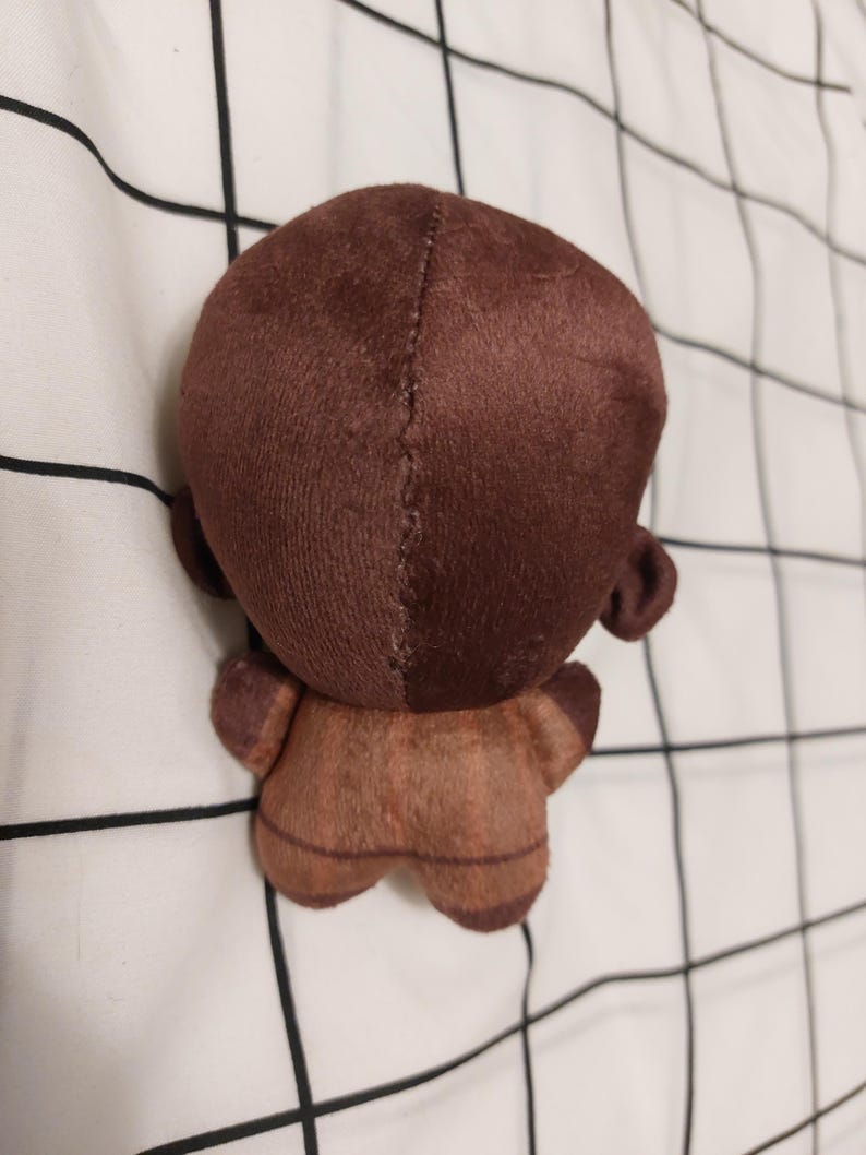 May include: A small, brown plush doll with a rounded head, small ears, and a stitched seam down the back. The doll's body is a lighter shade of brown, and it has small arms and legs.