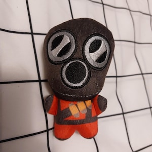 May include: A plush toy featuring a stylized character with a dark gray head and large, circular eyes. The character wears a red and gray outfit with orange and yellow accents. The toy is designed to resemble a gas mask.