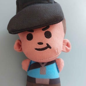 May include: A plush doll featuring a character with a peach-colored face, black embroidered eyes, and a small black smile. The doll wears a black cap, a bright blue shirt with a dark gray diagonal strap, and dark brown pants with a gray belt.