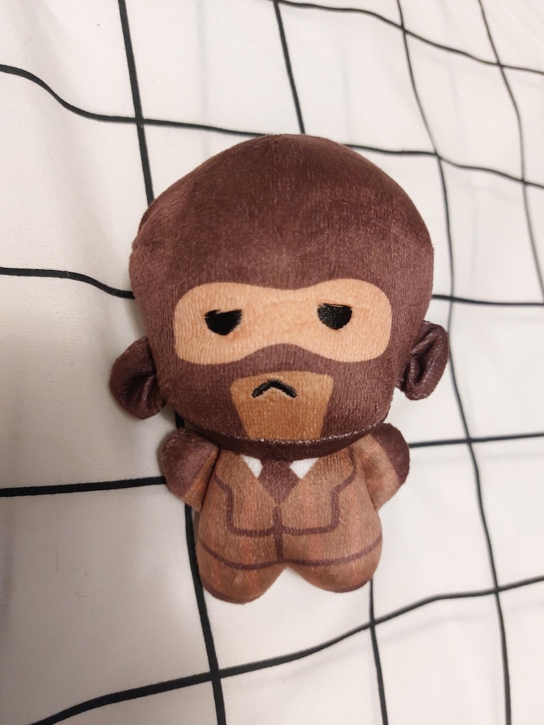 May include: A soft toy monkey wearing a brown suit and a mask. The monkey has a brown head and body with a tan face and a black mask. The toy is on a white surface with a black grid pattern.