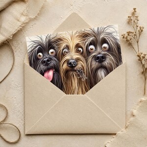 May include: A rectangular greeting card tucked inside a light tan envelope, featuring a detailed illustration of three shaggy dogs with wide eyes. The left dog is black with a pink tongue, the center is golden-brown, and the right is dark gray.
