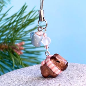 Puede incluir: Un amuleto de capibara marrón con una bufanda a rayas rosa y blanca, un amuleto de taza azul y blanca con lunares y cuentas transparentes y blancas que cuelgan de una cadena plateada.