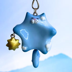 Puede incluir: Un dije en forma de gato azul claro con detalles blancos y un dije de estrella dorada. El gato tiene un lazo dorado en la parte superior para colgar. El dije tiene un acabado brillante y está suspendido sobre un fondo borroso.