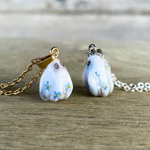 Può includere: Due delicate collane con un ciondolo in porcellana bianca decorato con fiori blu di nontiscordardime. Una collana ha una catena dorata e l'altra una catena argentata.