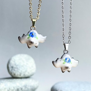 Peut inclure: Deux chaînes en argent avec des pendentifs en forme de chauve-souris en céramique blanche avec des accents de fleurs bleues. La chauve-souris a une petite bouche noire et des yeux.