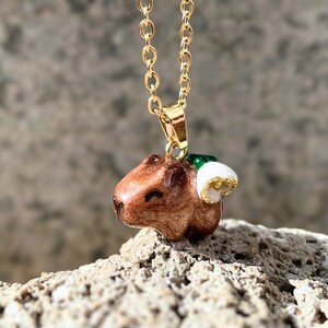 Può includere: Una collana con catena dorata con un ciondolo capibara marrone e bianco. Il capybara indossa un cappello verde e bianco.