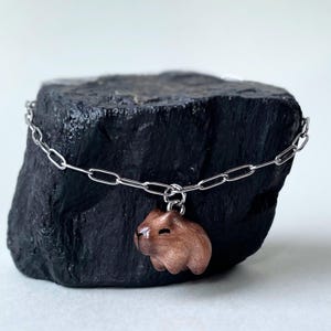 Peut inclure: Un bracelet à chaîne argentée avec un charm en forme de capybara marron. Le charm est une sculpture tridimensionnelle détaillée de l'animal. Le bracelet est présenté sur une surface sombre et texturée, créant un contraste.