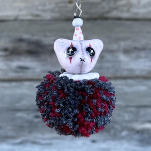May include: A handmade clown-themed keyring. The clown's head is pale pink with black cross-shaped eyes, red tear streaks, and a black 'X' mouth. It has a white collar and a red and black pom-pom body. A small pink party hat with white polka dots sits atop the head.